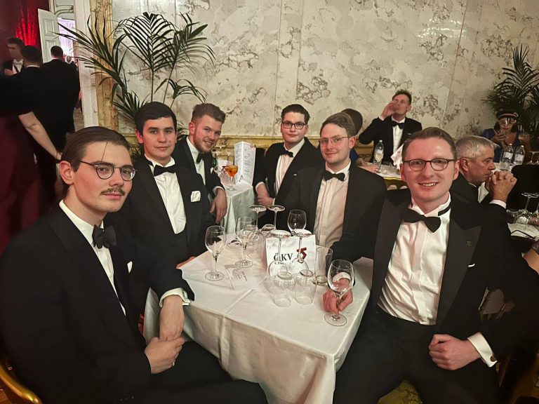 Sechs lächende, junge Männer im Smoking sitzen um einen kleinen Tisch mit Weingläsern. Andere sehr schick gekleidete Menschen sitzen und gehen im hintergrund.