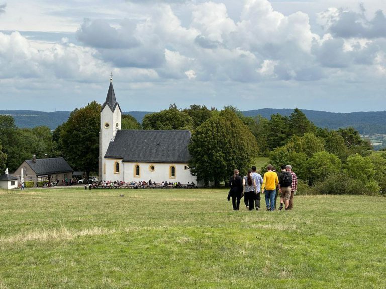Eine Gruppe von sechs Menschen von hinten gesehen läuft über eine große Wiese auf eine Kirche vor einer weiten Landschaft zu.